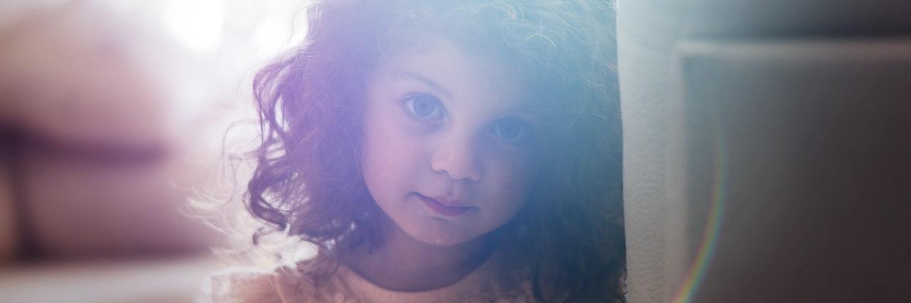 Young girl with curly brown hair wearing an orange top leans against a wall and looks directly at the camera