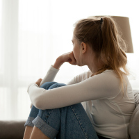photo of young woman sitting on couch looking away from camera toward window, resting chin on her hand
