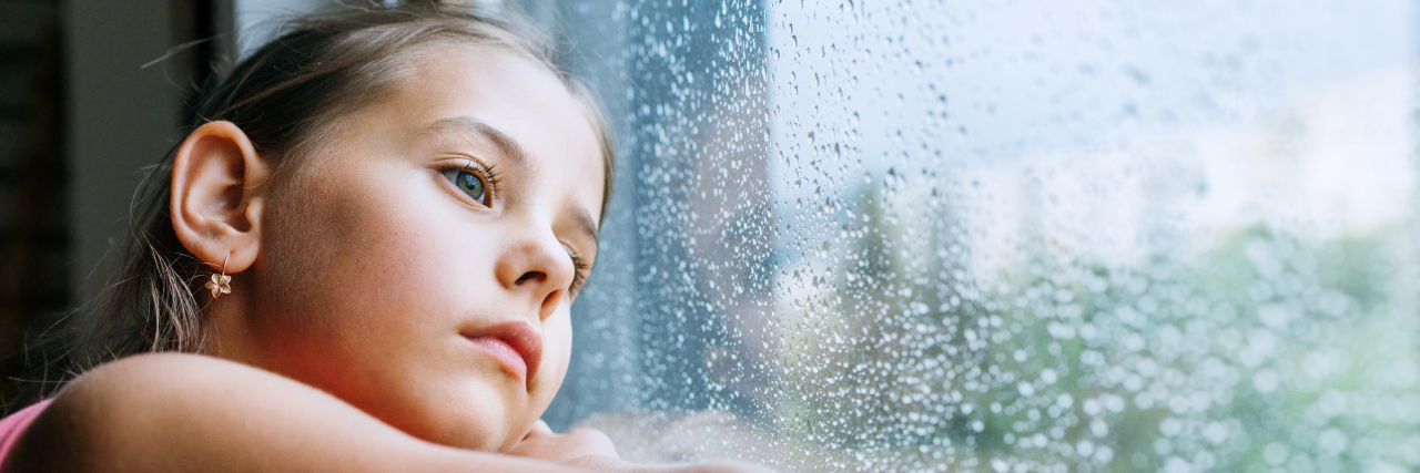 Little sad girl looking through the window on a rainy day.