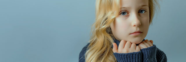A young girl resting her head on her hands, looking serious and straight ahead. She has long blonde hair and blue eyes.