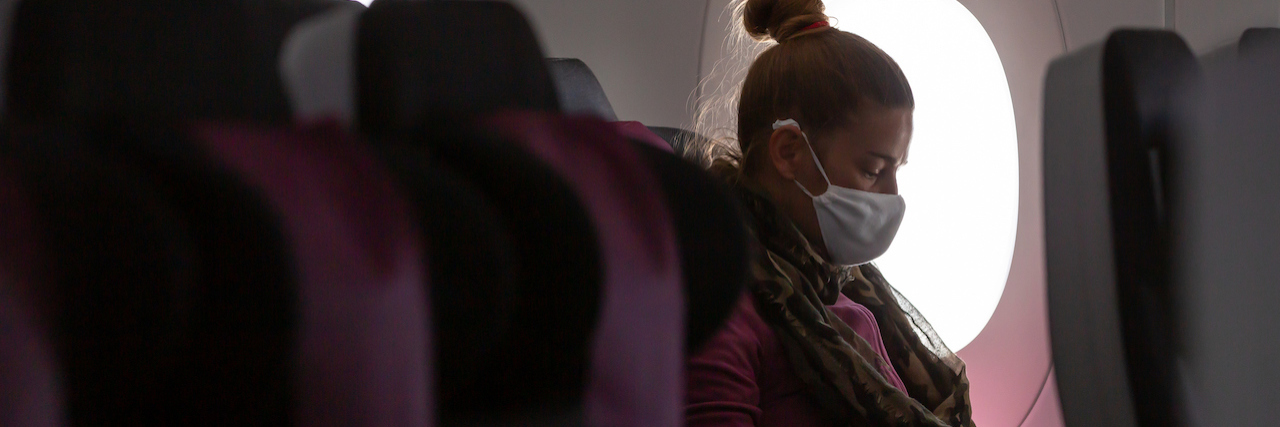 What Is the Risk of Catching the Coronavirus on a Plane? A woman sits in the window seat of an airplane with her hair in a bun wearing a face mask and gloves