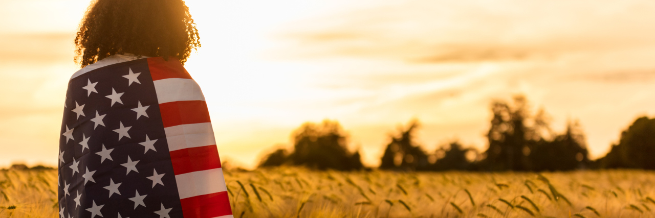 black woman standing in field with American flag wrapped around her, looking at sunset