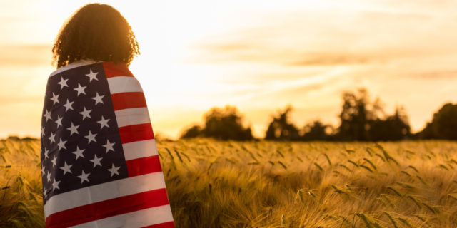 black woman standing in field with American flag wrapped around her, looking at sunset