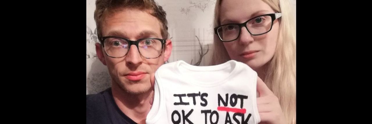a couple holding a baby onesie that says "it's not OK to ask when"