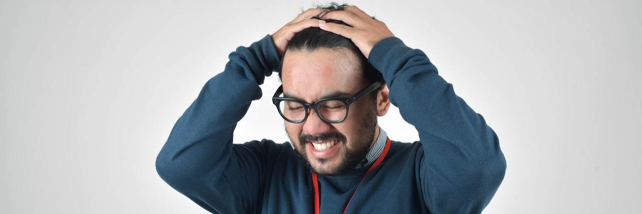 Frustrated-looking man with dark hair and glasses with his hands on his head