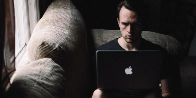 photo of man sitting on sofa beside a window with a laptop on his knees
