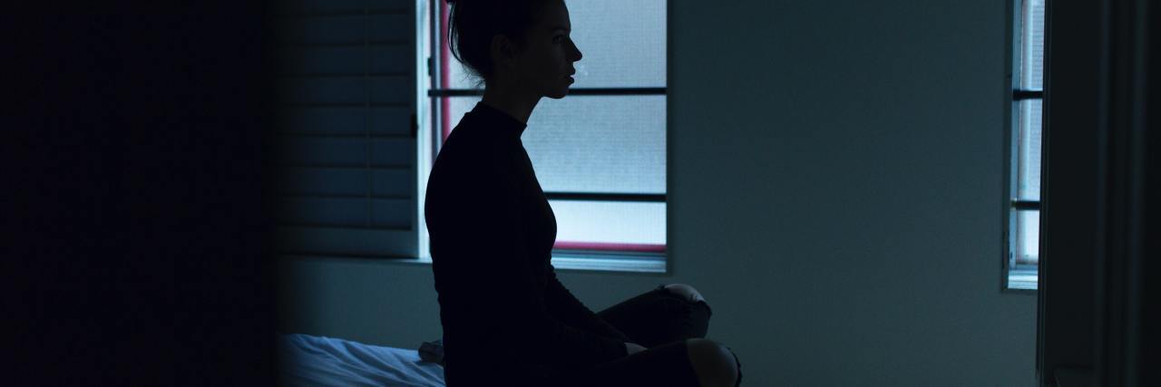 I Don't Restrict but That Doesn't Mean I Don't Have an Eating Disorder photo of a woman silhouetted against window while sitting on bed in bare room