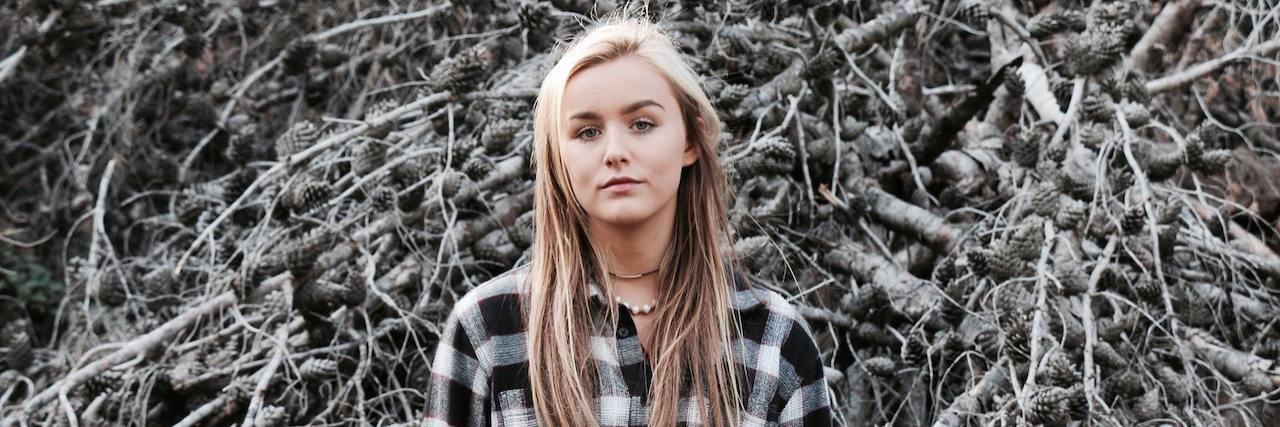 How to Help Someone Who Is Suicidal from Attempt Survivors Woman wearing an oversized black and white plaid shirt standing in front of a leaf-less bush