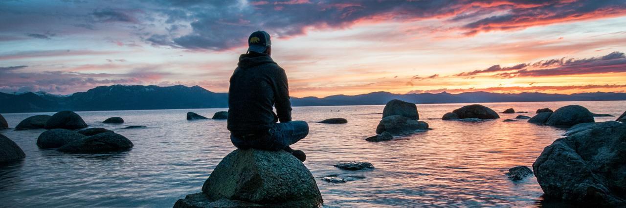 photo of man sitting on a rock by the ocean at sunset