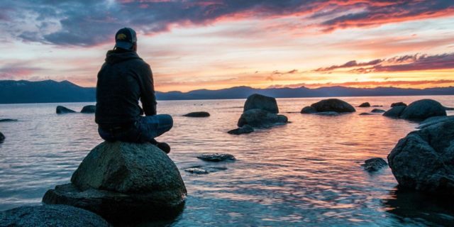 photo of man sitting on a rock by the ocean at sunset