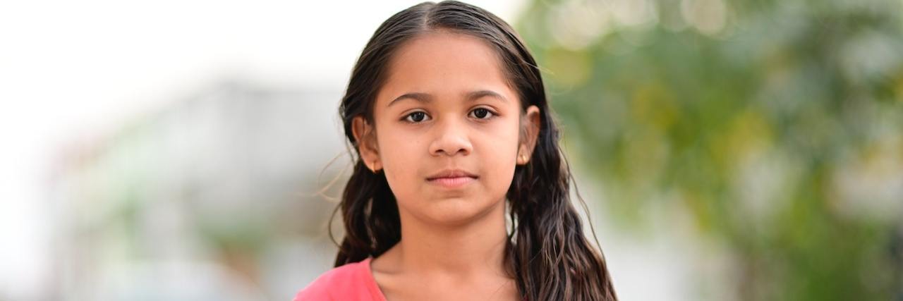 What It Sounds Like When Your Parent Is a Gaslighter Young girl with long dark hair wearing a pink shirt and standing outside looking directly at the camera