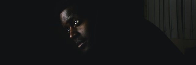 photo of black man sitting in darkness, looking up, with tears in his eyes