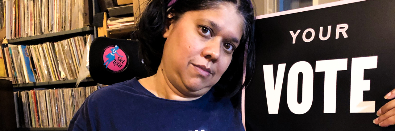 How People With Disabilities Are Excluded From Voting and Activism Priya sitting next to a sign that says "vote."