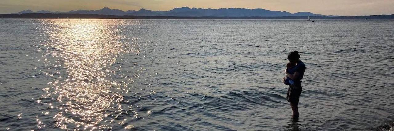 Proactively Supporting My Child's Mental Health Woman holding infant son, standing in shallow water at the beach at sunset
