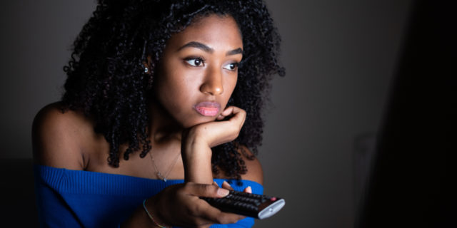 close-up of a young Black woman holding a remote control, pointed at a tv