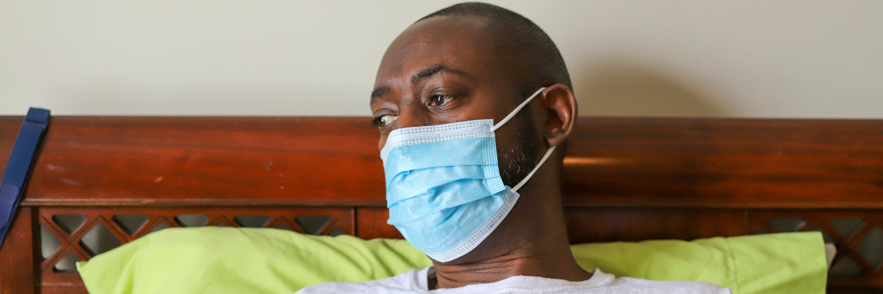 A man wearing a protective face mask in bed while looking toward a window