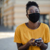 Black woman in a yellow shirt, wearing a mask while smiling at the camera, phone in hand