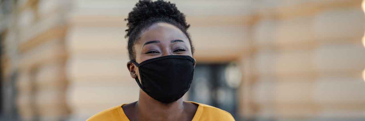 7 Ways to Create Joy During COVID-19 and Other Uncertain Times Black woman in a yellow shirt, wearing a mask while smiling at the camera, phone in hand