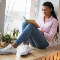Young Black woman sitting in the window reading a book.