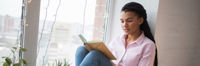 Young Black woman sitting in the window reading a book.