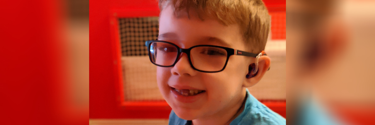 Why Awareness Days Matter to Me as a Parent of a Kid With a Rare Disease Liam Ferguson, a young boy with blonde hair, glasses and a hearing aid, wearing a blue shirt