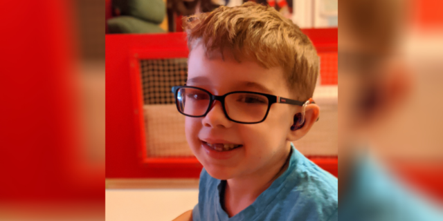 Liam Ferguson, a young boy with blonde hair, glasses and a hearing aid, wearing a blue shirt