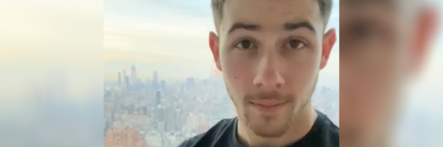 Nick Jonas standing in front of a window that shows a hazy city skyline