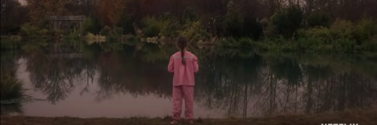 Young girl wearing pink pajamas looks out over a creepy lake surrounded by trees