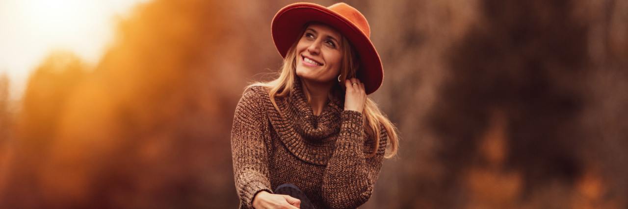Lessons Learned After Decades of Living With Bipolar Disorder photo of a happy older woman sitting on the ground at sunset, wearing a hat, with trees in the background