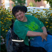 Chelsie smiling as she sits in her power wheelchair outdoors with flowers in the background.