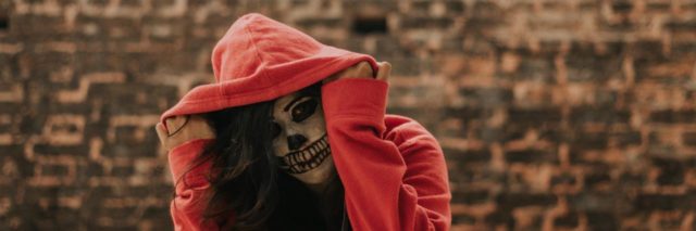 photo of person with red hooded sweatshirt pulled over head, wearing skull makeup for Halloween, against brown brick wall with bars over window