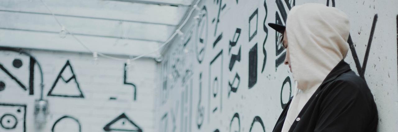 Listen When People With Mental Illness Tell You What They Need Man sitting in a white room with symbols on the walls wearing a white hoodie over his head, black jacket, jeans and black boots