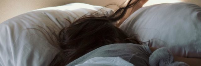 Person with long brown hair lying with their face hidden in a pillow in bed