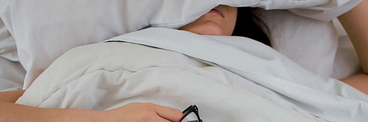 What to Know About Buying a Weighted Blanket for Mental Health photo of woman under a blanket and covering her face with a pillow while holding a pair of glasses