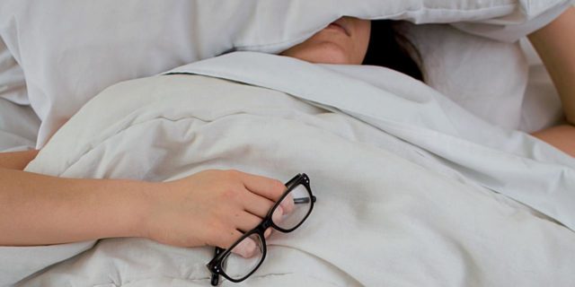 photo of woman under a blanket and covering her face with a pillow while holding a pair of glasses