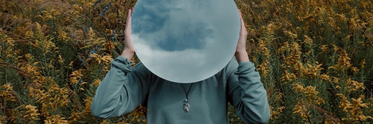 A woman holding a mirror over her face standing in the field of flowers. The mirror is reflecting a cloudy sky