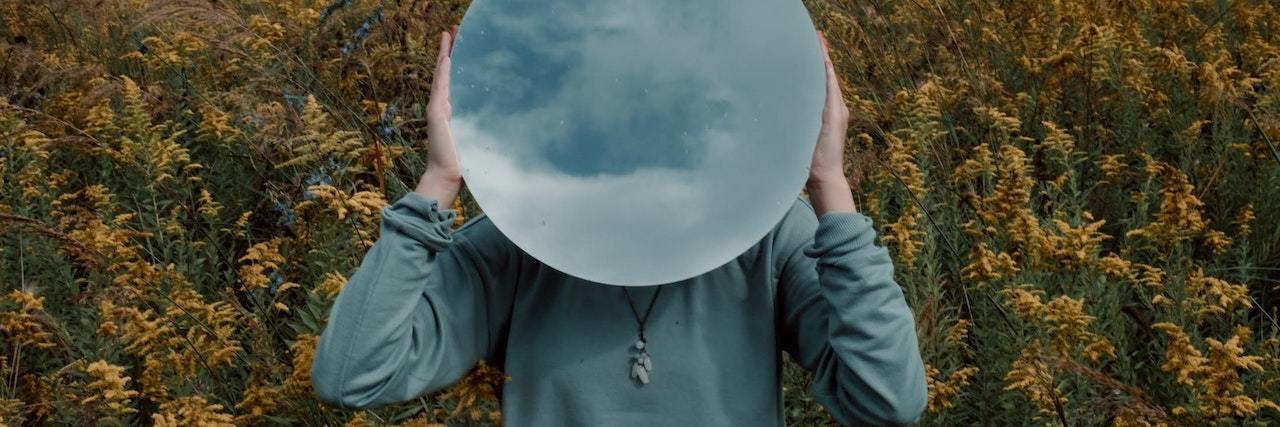 The Void: Description of What Depression Feels Like A woman holding a mirror over her face standing in the field of flowers. The mirror is reflecting a cloudy sky
