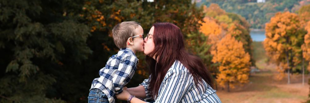 The Loneliness of Parenting a Child With Cerebral Palsy Kristina giving her son a kiss outside with autumn trees in background.