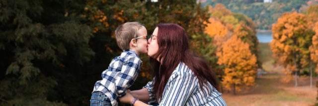 Kristina giving her son a kiss outside with autumn trees in background.