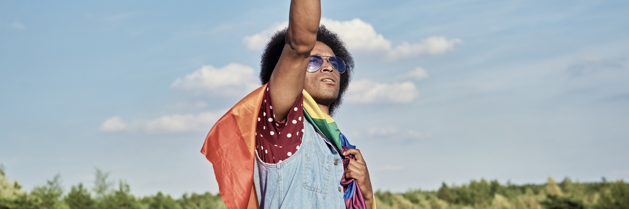 photo of man with a rainbow flag, raising his fist to the blue sky