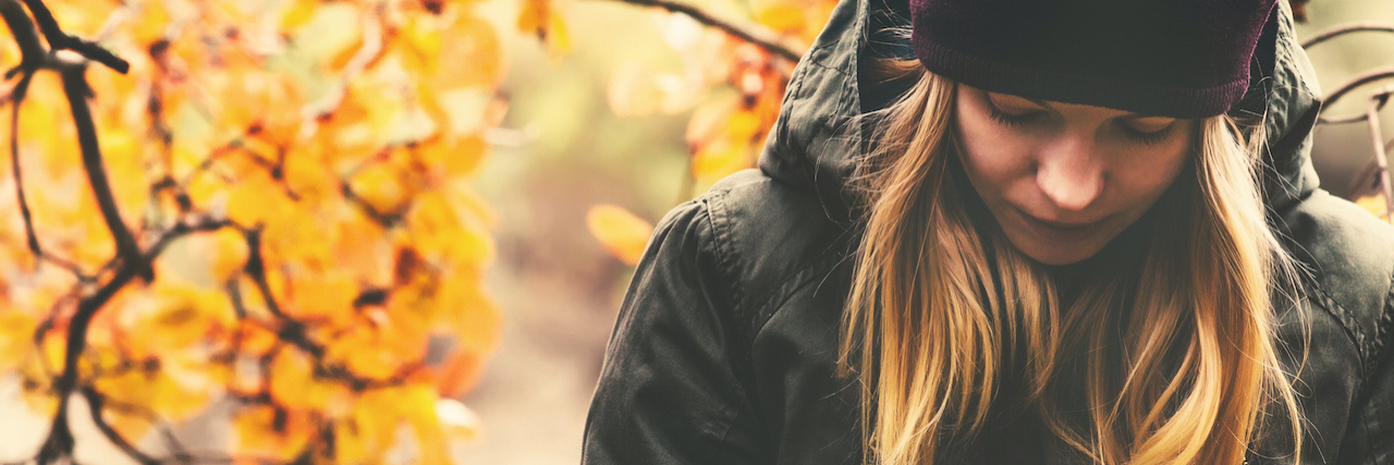 Experiencing Complex Grief During the Holidays woman next to fall leaves, wearing a coat and a hat, looking down