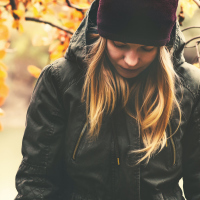 woman next to fall leaves, wearing a coat and a hat, looking down