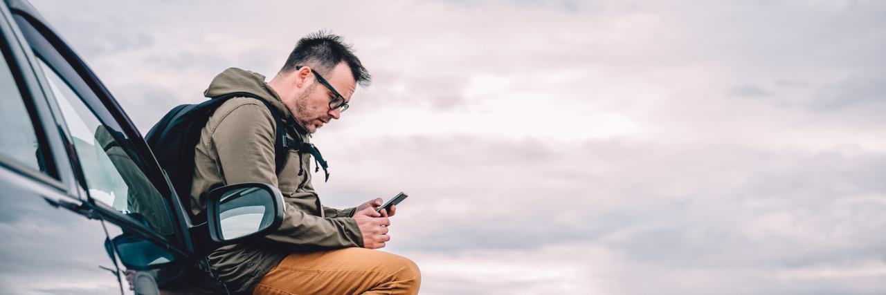 My Dead Car Battery Taught Me to Stop Isolating With Schizophrenia White man sitting on his car, texting someone, on a deserted road