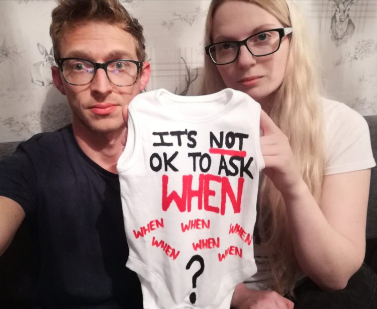 a couple holding a baby onesie that says "it's not OK to ask when"