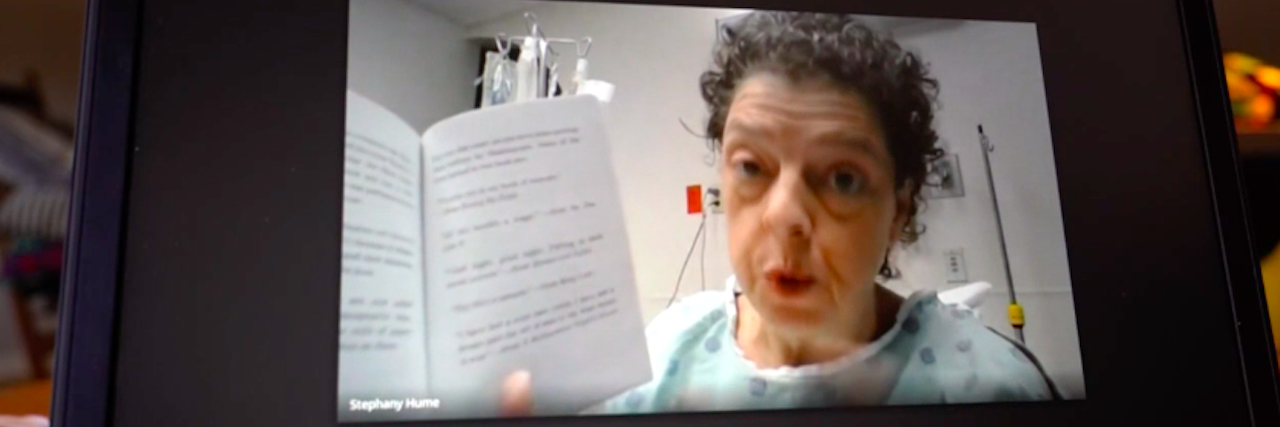 Stephany Hume, a female teacher with short dark gray hair, wears a hospital gown and holds a book up to the camera of a video conference screen