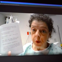 Stephany Hume, a female teacher with short dark gray hair, wears a hospital gown and holds a book up to the camera of a video conference screen