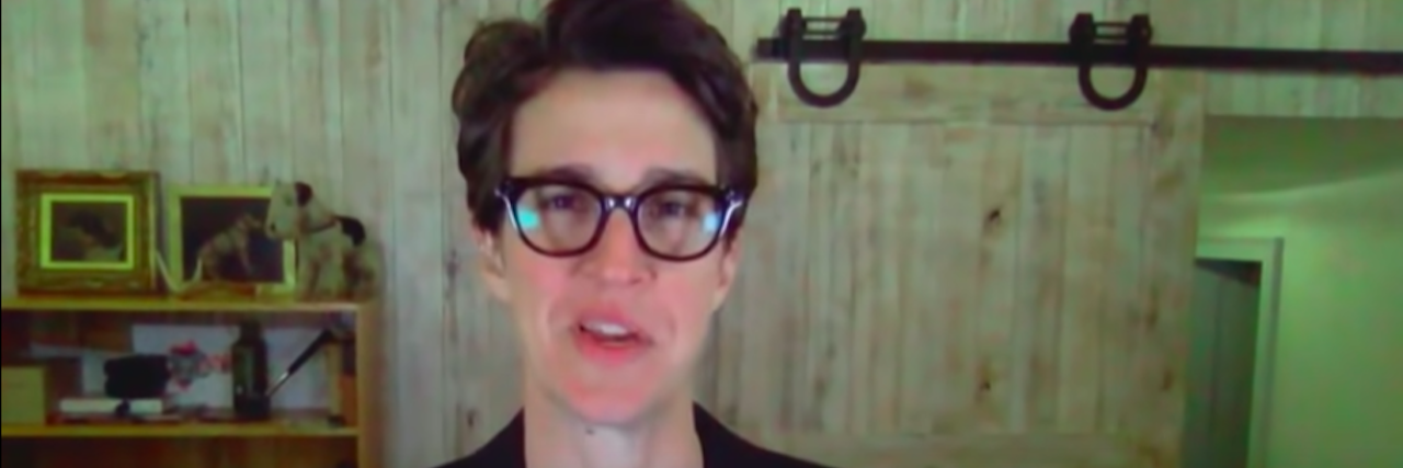 Rachel Maddow, a woman with shirt brown hair glasses and a dark suit, speaks to the camera at her home