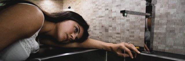 photo of woman resting her head beside a sink with water running
