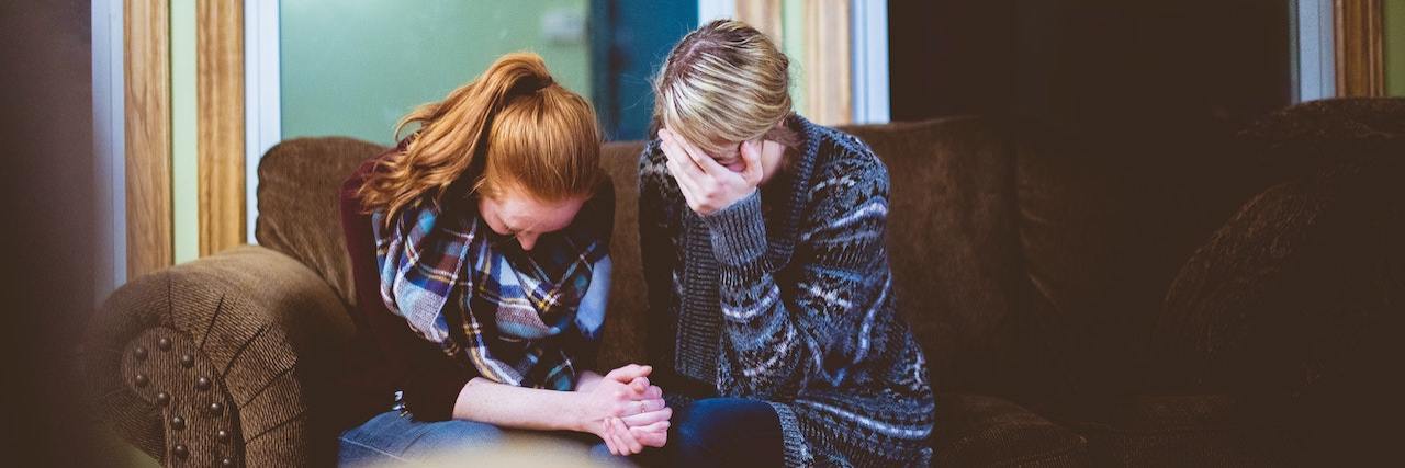 6 Things to Know If Someone You Love Struggles With Addiction Two women sit side by side on a couch with their heads down looking distressed and holding hands