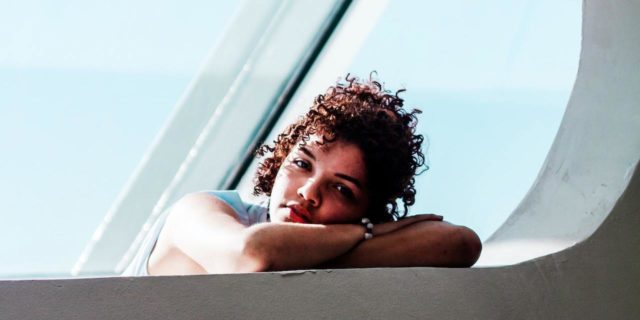 Sad-looking woman with curly brown hair resting her head on her arms in front of a window